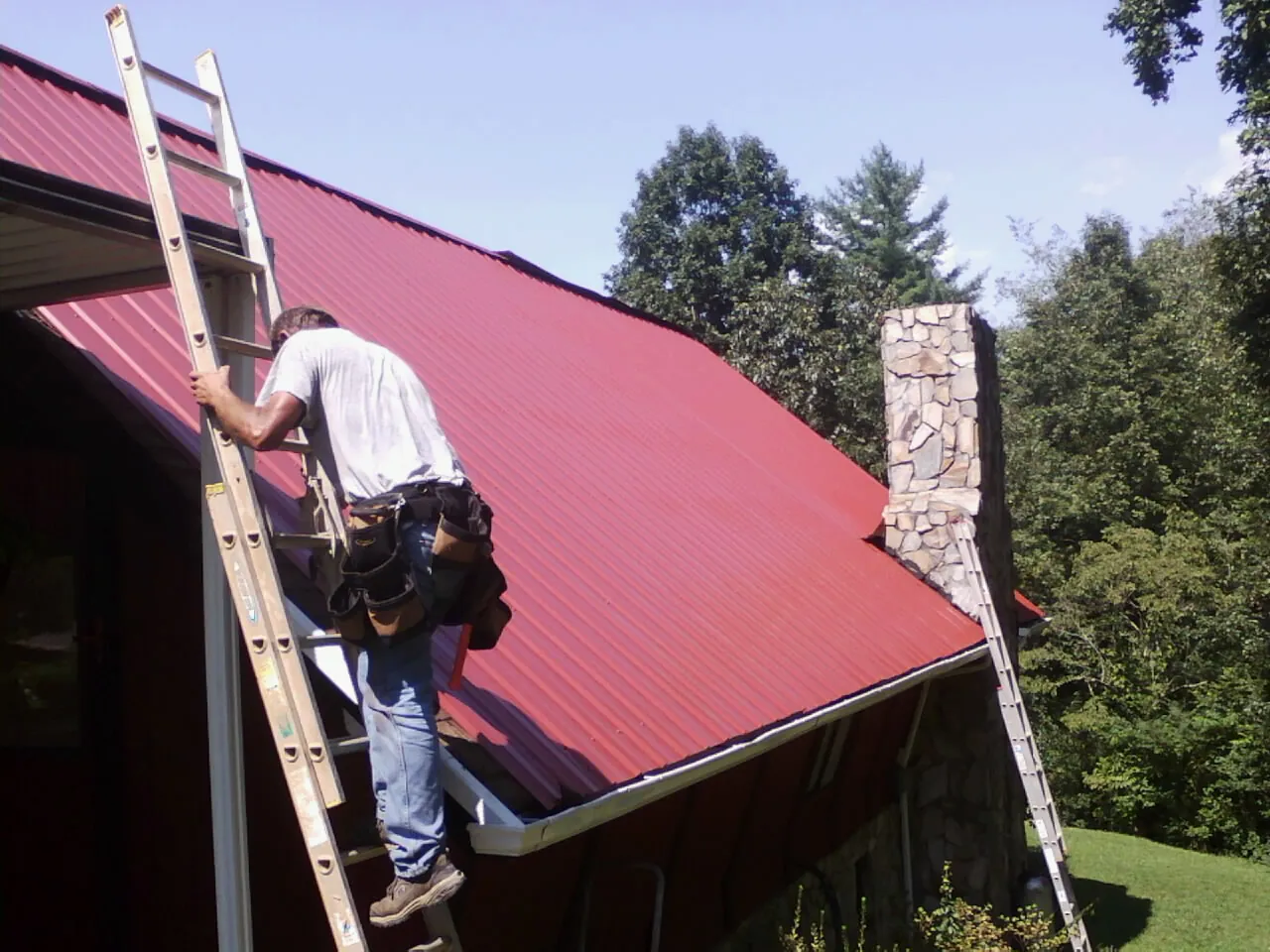 Roofer on a ladder beside a red metal roof and stone chimney — Soffit & Fascia Repair by Roofing Brothers in Cosby